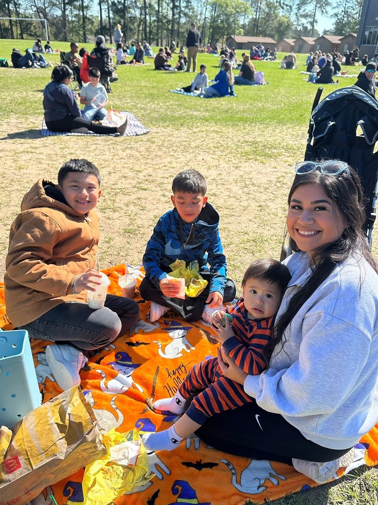 Family enjoying a picnic on a Halloween-themed blanket in a sunny park with other groups in the background.