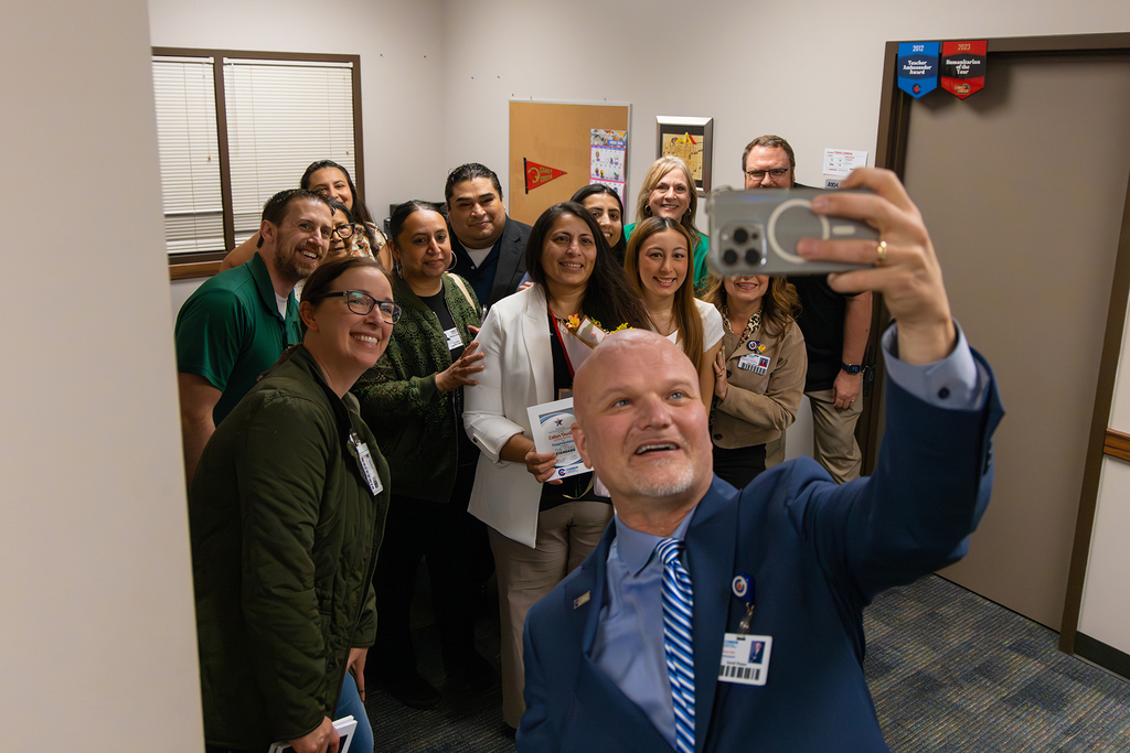 Group of staff gather for selfie in office as administrator holds phone.
