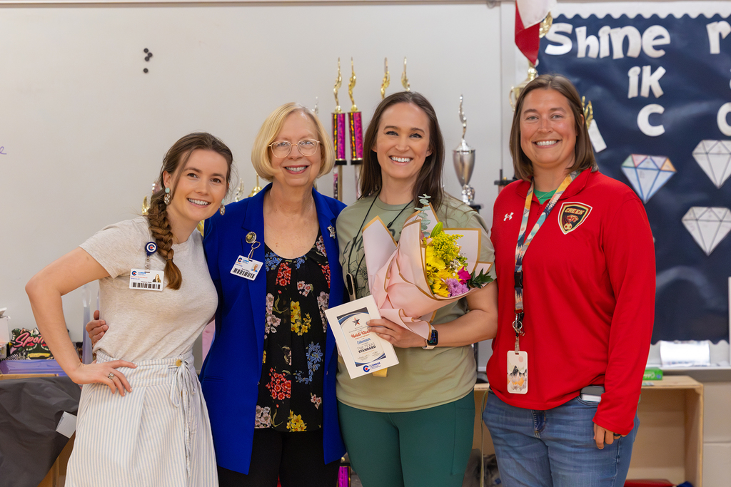 Four educators pose in classroom with teacher holding flowers and recognition plaque.