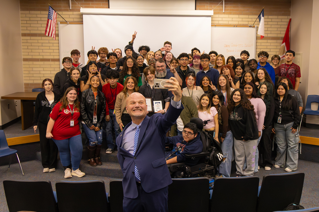 Administrator takes selfie with large group of students and staff in classroom.