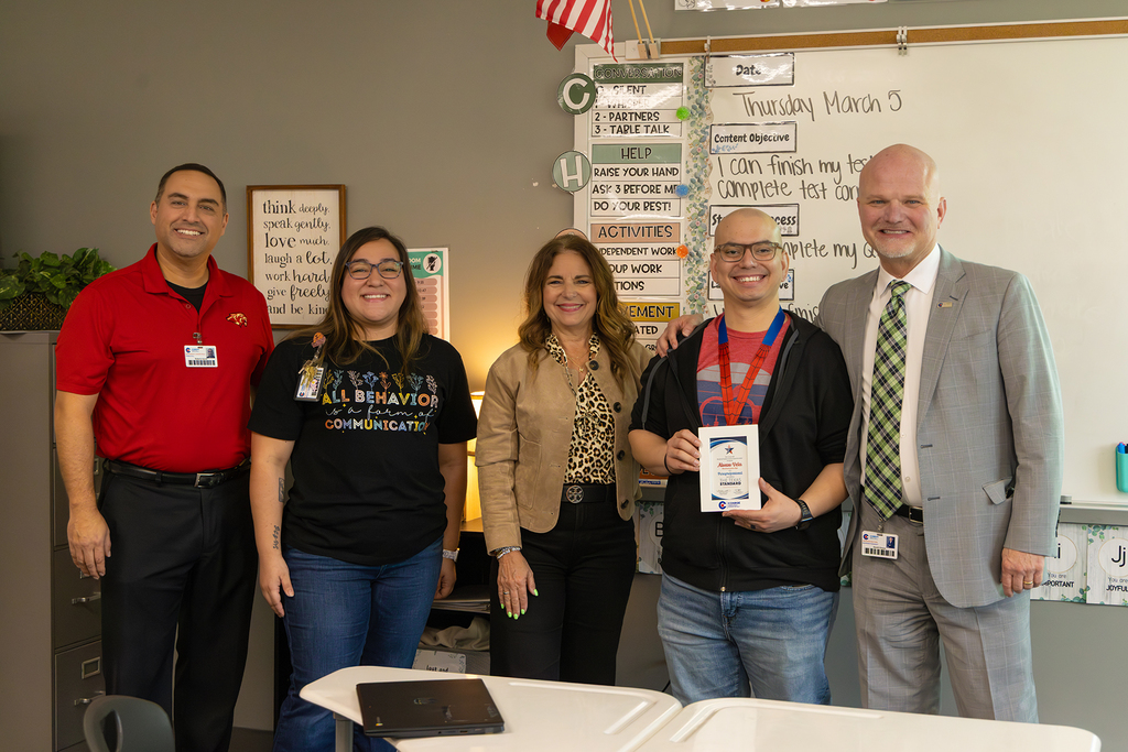 Group of educators pose in classroom with teacher holding recognition plaque.