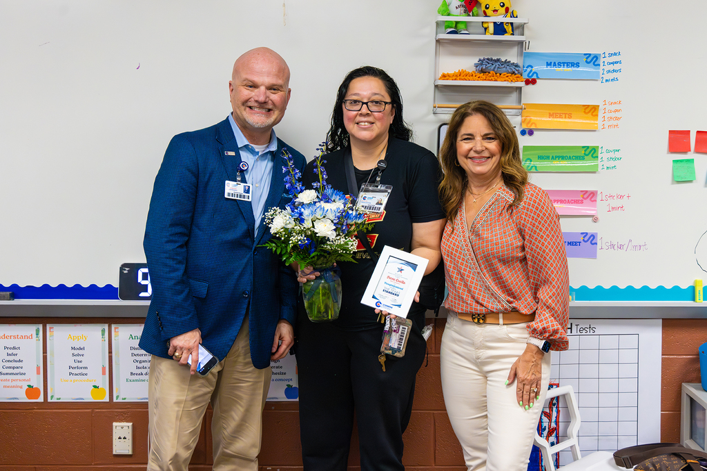 Superintendent and board member in classroom with educator holding flowers and award.