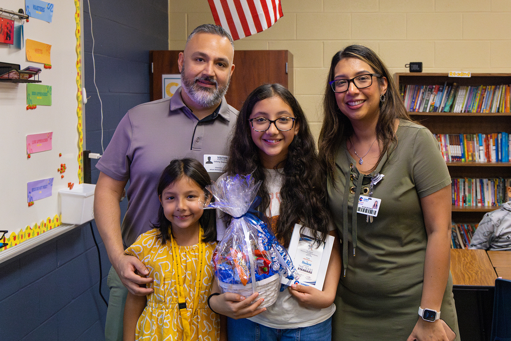 Family and staff pose in classroom with student holding gift basket and recognition plaque.