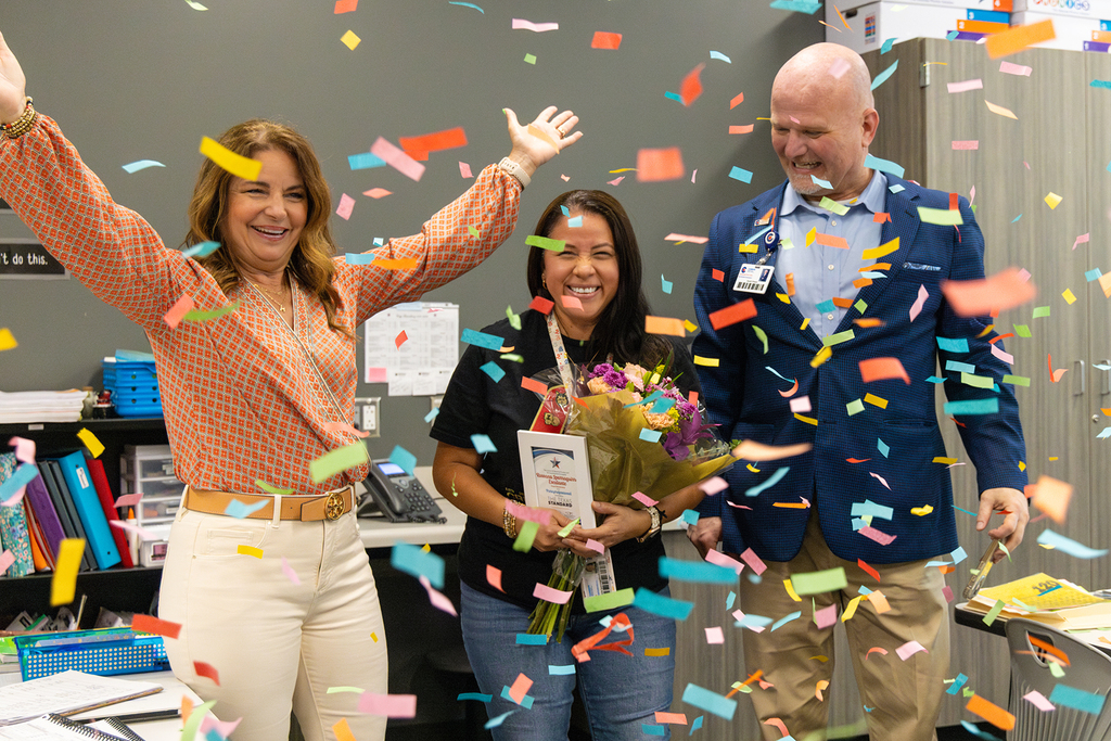 Board member and superintendent celebrate educator with confetti as she holds flowers and recognition plaque.