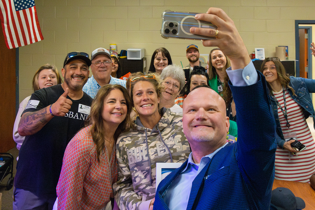 Group of staff gather for selfie in classroom as administrator holds phone.
