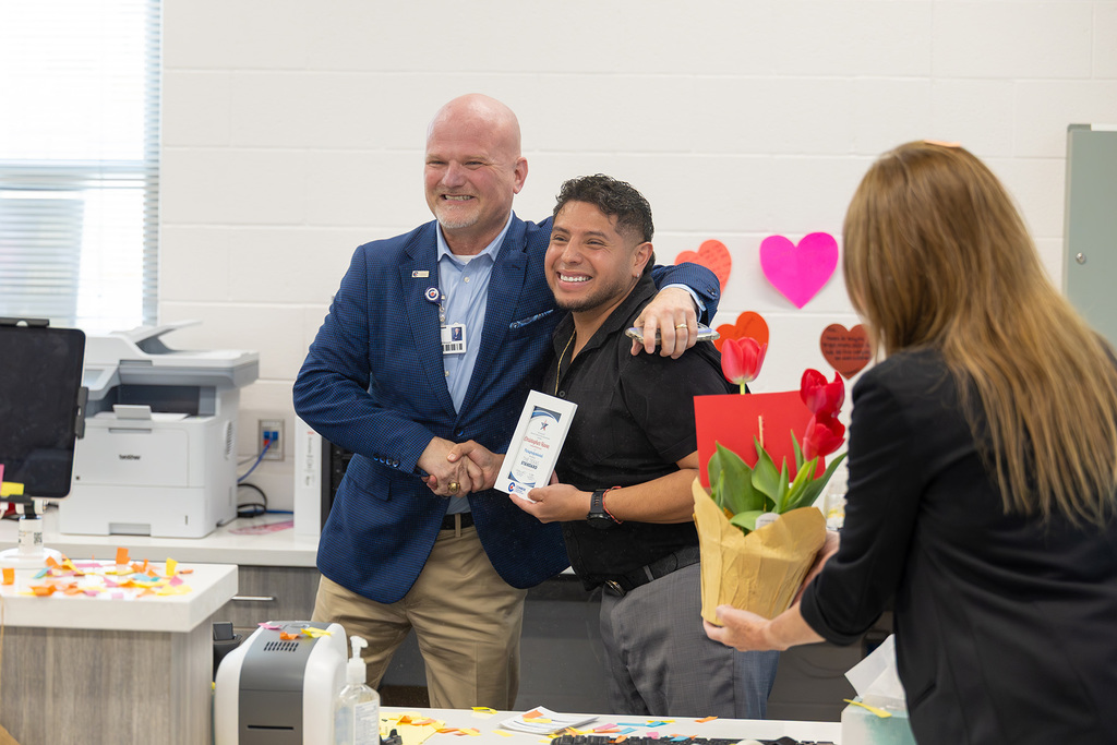 Superintendent congratulates staff member with handshake and award in office.