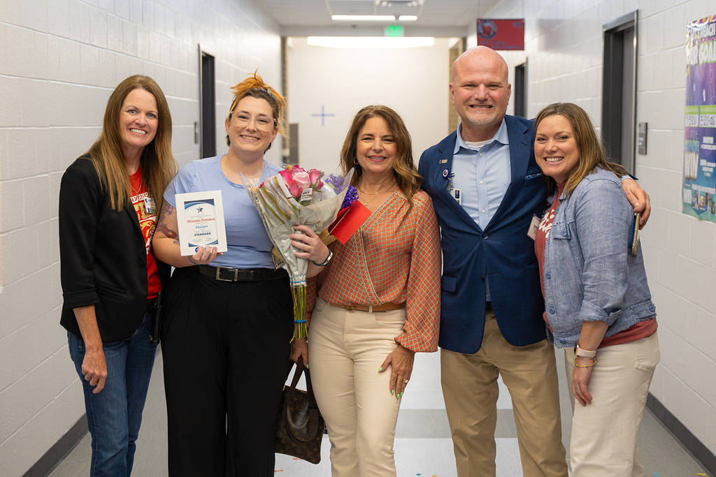Staff pose in hallway with educator holding flowers and recognition certificate.