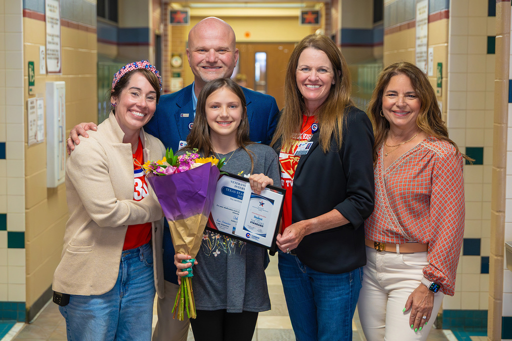 Student stands with staff in hallway holding flowers and recognition certificate.