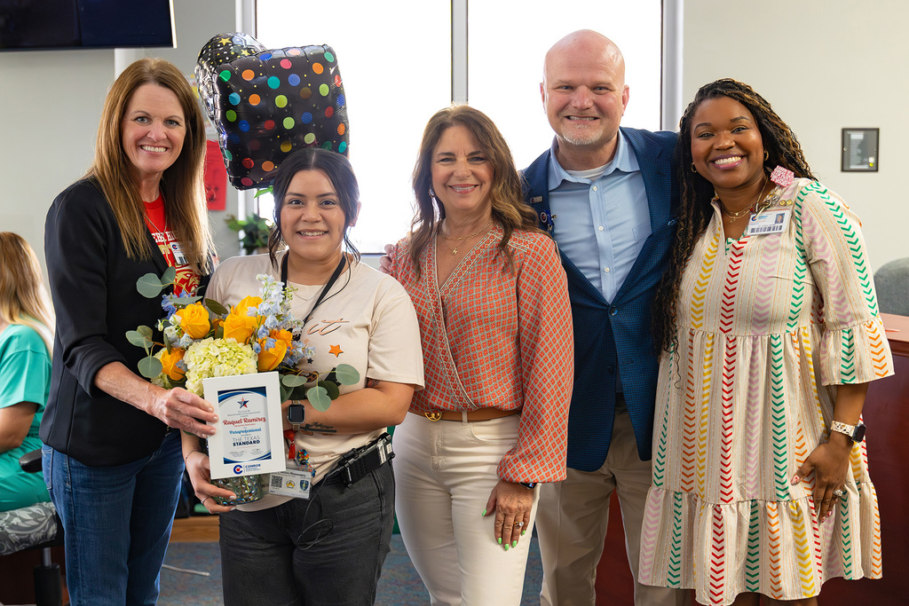 Five staff members pose indoors with educator holding flowers and recognition plaque.