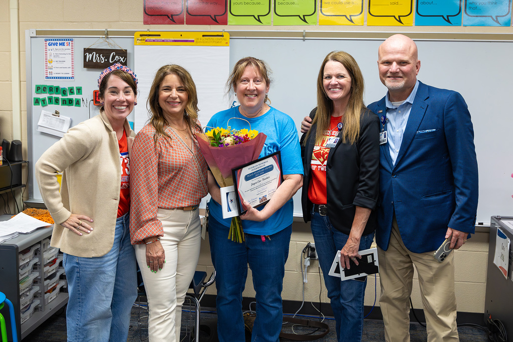 Group of staff pose in classroom with educator holding flowers and framed award.