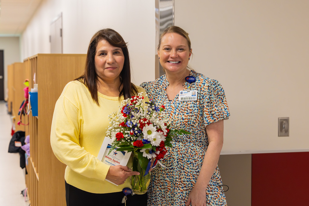 Principal and educator stand together in hallway with flowers and recognition certificate.