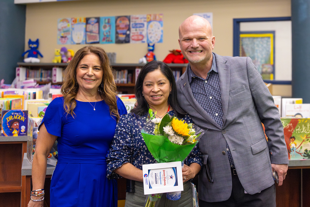 Board member and superintendent pose in library with educator holding flowers and award.