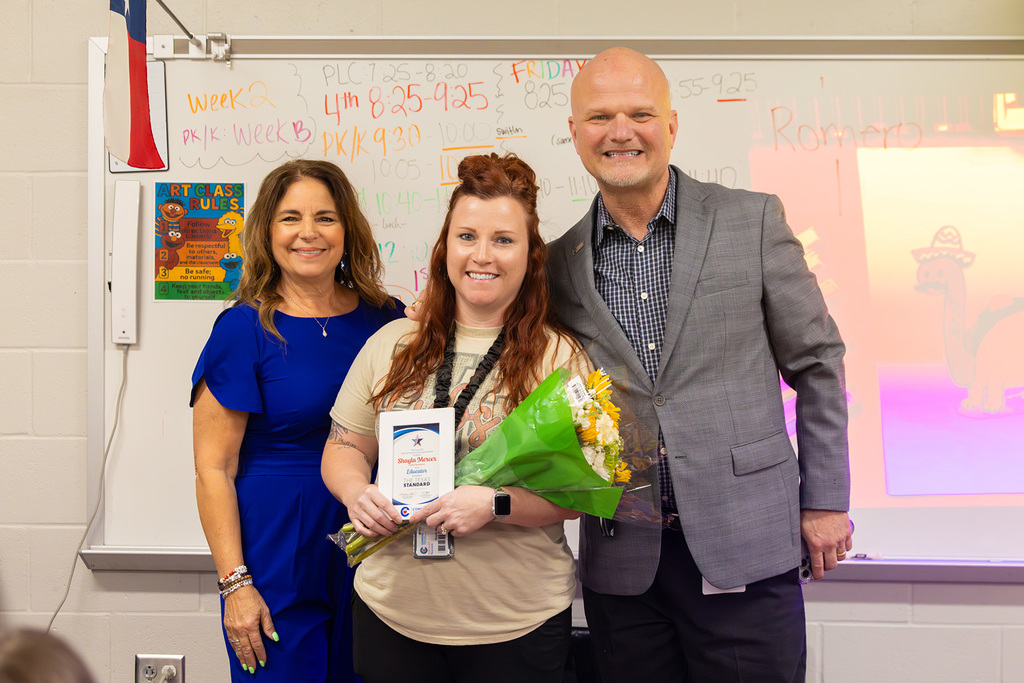Board member and superintendent pose in classroom with educator holding flowers and award.