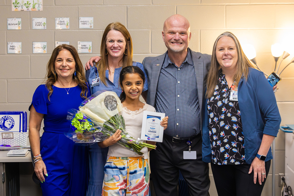 Student stands with staff holding flowers and recognition certificate.