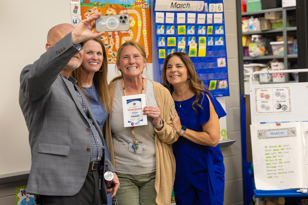 Superintendent and board members take a selfie with teacher holding recognition certificate in classroom.