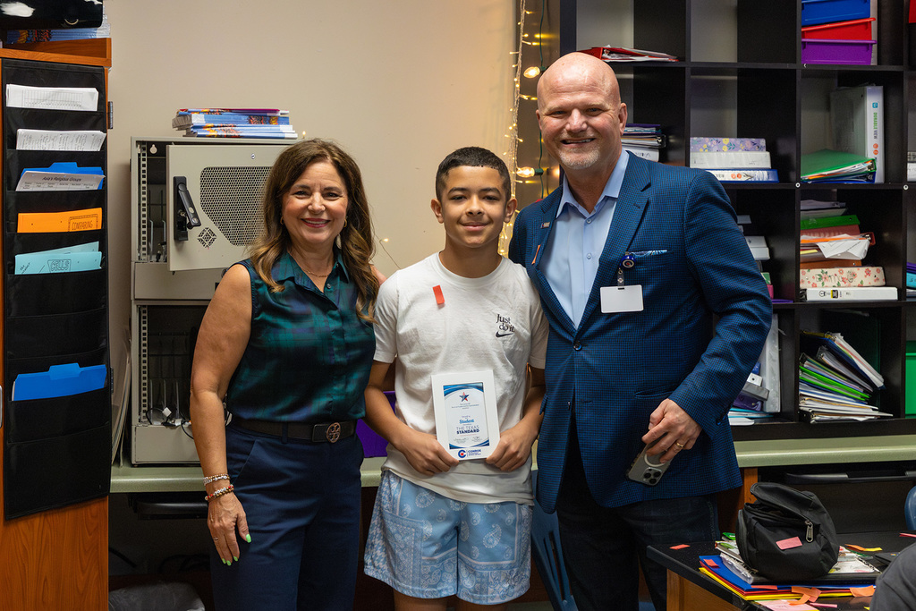 Student stands with board member and superintendent holding recognition certificate in classroom.