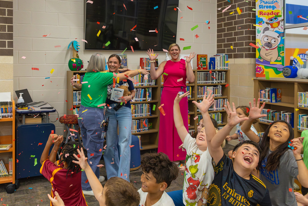 Students celebrate with confetti as staff hug award recipient in library.