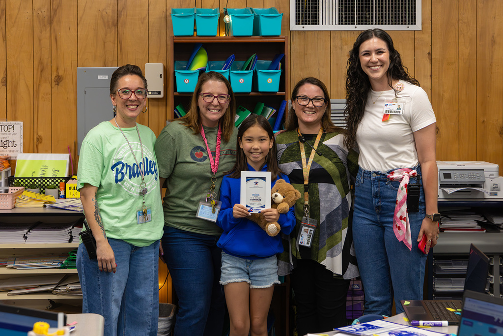 Student stands with staff holding certificate and teddy bear in classroom.