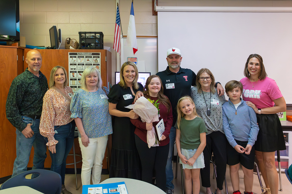 Group of family and staff pose with student holding flowers in classroom.