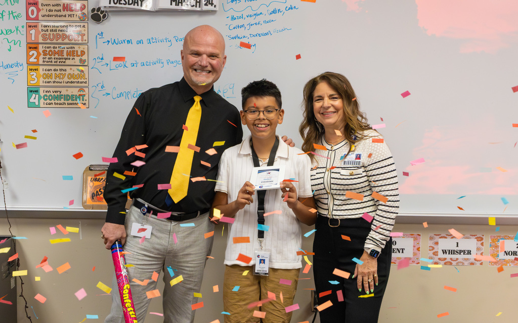 Student holding a certificate stands between two adults in a classroom with colorful confetti falling around them.