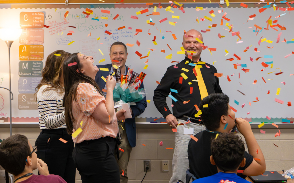 In a classroom, students and a teacher express excitement as bright confetti floats through the air
