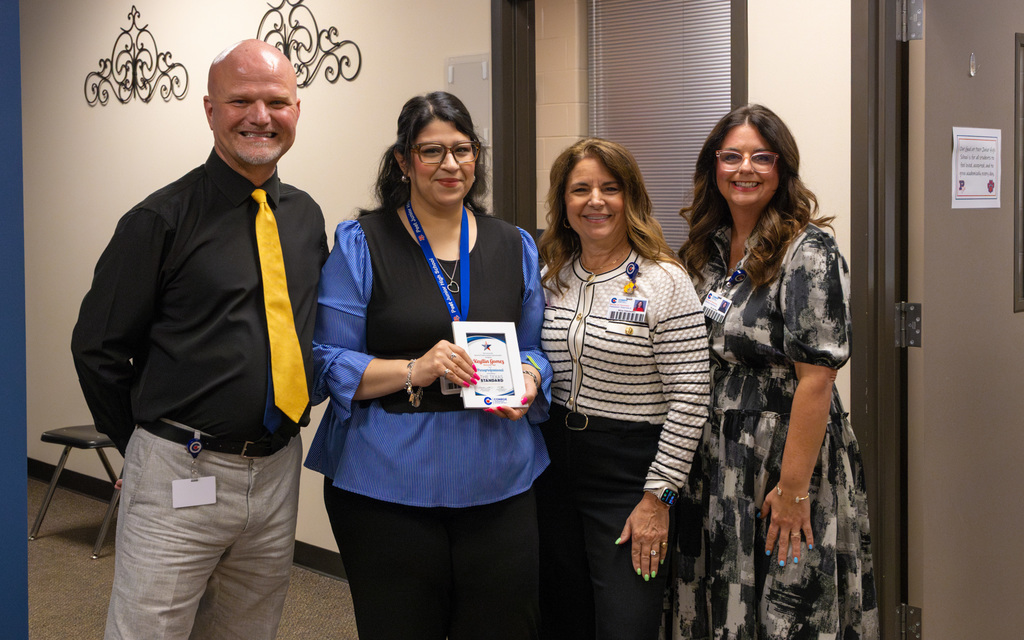 Four individuals standing together in an office setting, one holding a small framed certificate