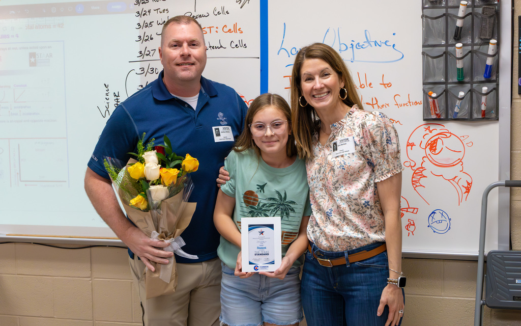 A family photo in a classroom where a girl holds a small framed certificate, a man holds flowers, and a woman stands beside them