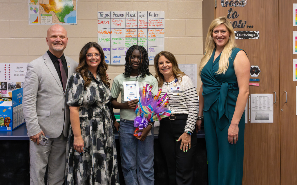 Classroom scene with five individuals, including a student in the center holding an award and a bright candy bouquet