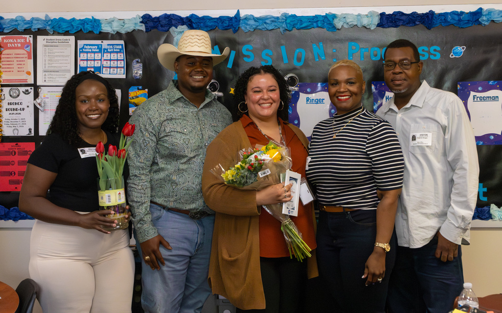 Five individuals standing together in a classroom, two holding bouquets of flowers and smiling for the photo