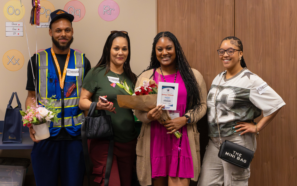 Group of four adults posing together, one in a safety vest, two holding flowers, standing in front of a wooden storage cabinet