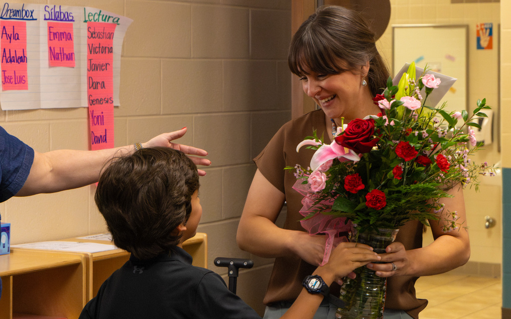 Staff member holding a mixed bouquet of red and pink flowers while interacting with a child in school