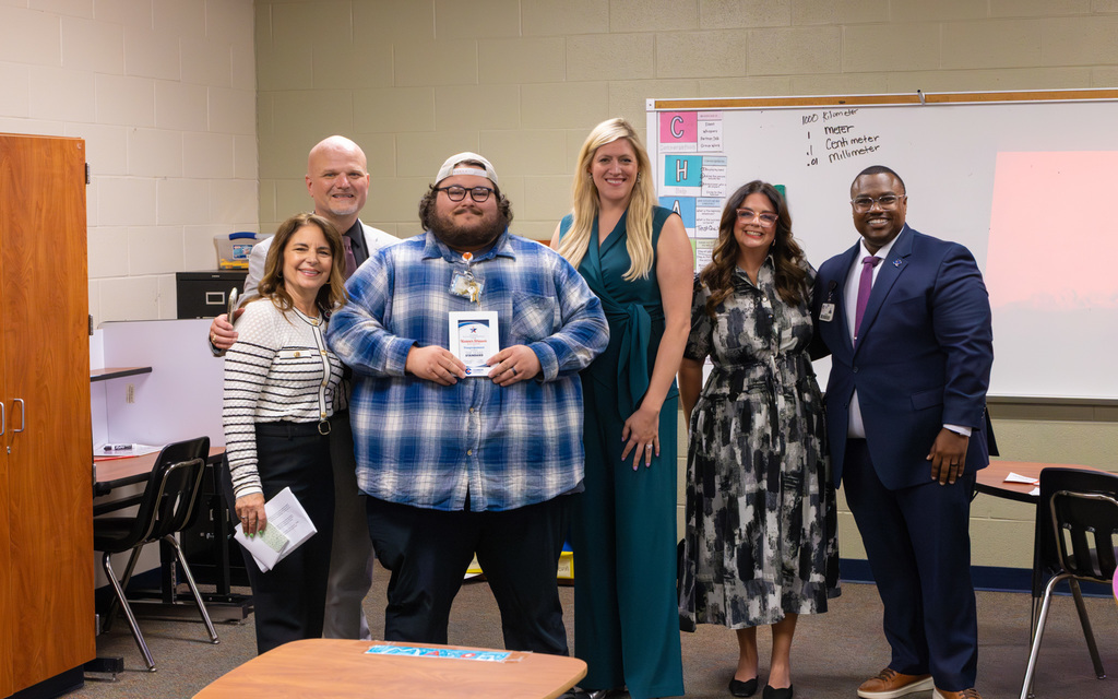 Group of six adults standing together in a classroom, including a person holding a certificate, all smiling for the photo
