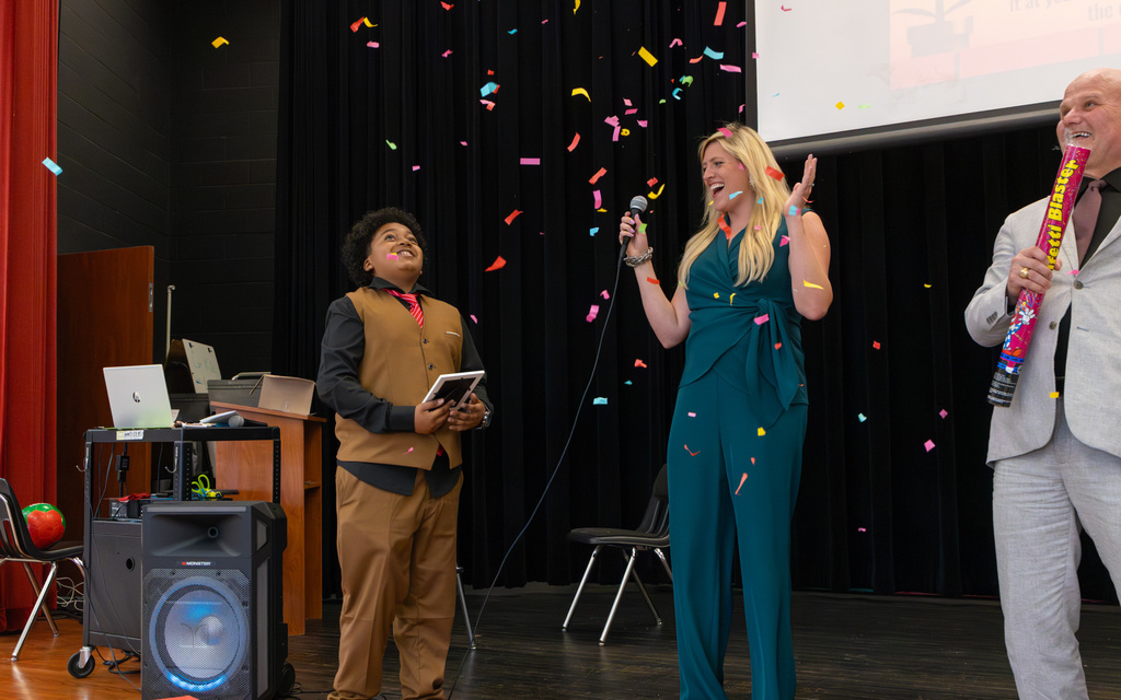 Lindsay Dawson, in a teal jumpsuit, holding a microphone on stage, celebrating with confetti, joined by Dr. Vinson and a student, both dressed in formal wear
