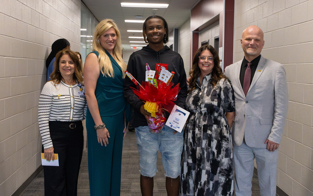 Five individuals stand in a hallway for a photo with a student in the center holding a framed award and a candy bouquet