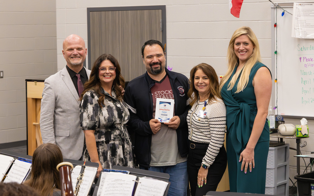 A group photo of five adults in a music room, including a person displaying a recognition award