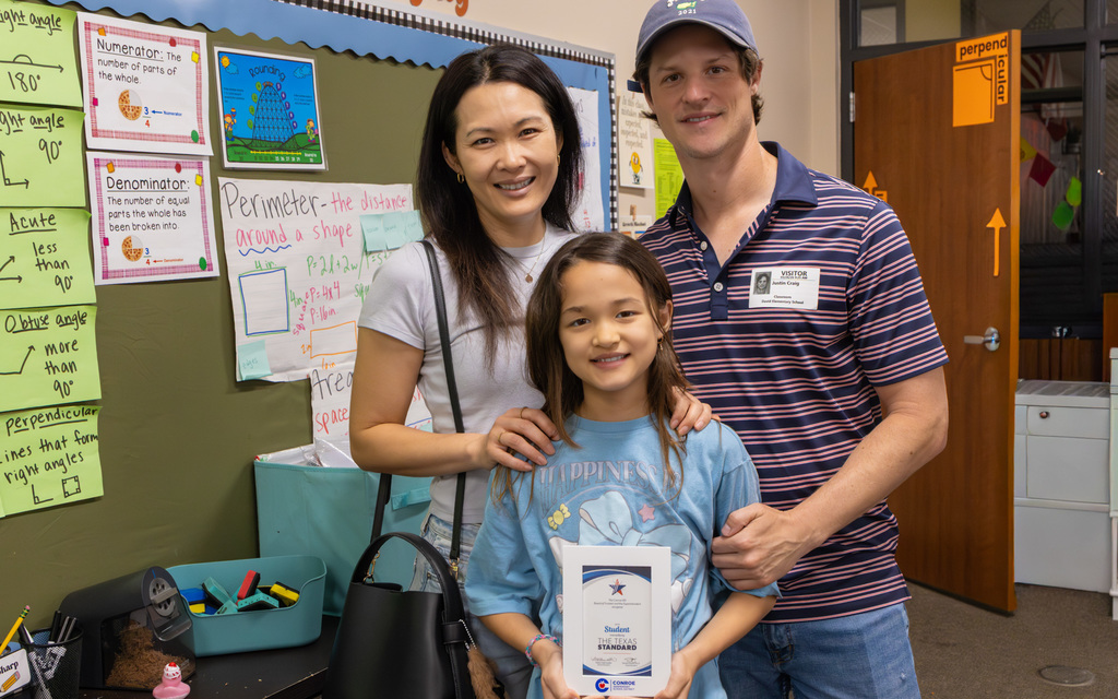 Family of three posing in a classroom with math posters on the wall and a student holding a certificate