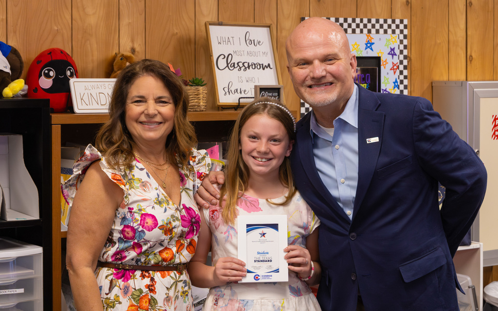 Girl holding a recognition certificate posing with two adults in a classroom featuring motivational signs and shelves