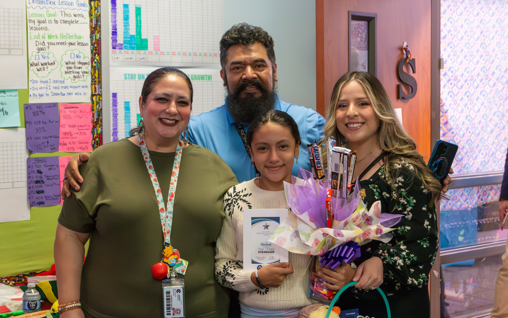 A family photo posing in a classroom setting, with a student holding an award and a decorative candy bouquet