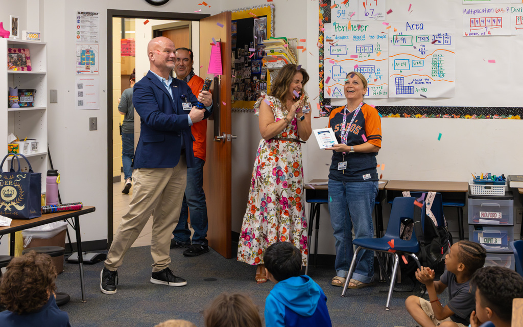 Classroom scene showing a teacher being recognized with an award by two adults while peers observe