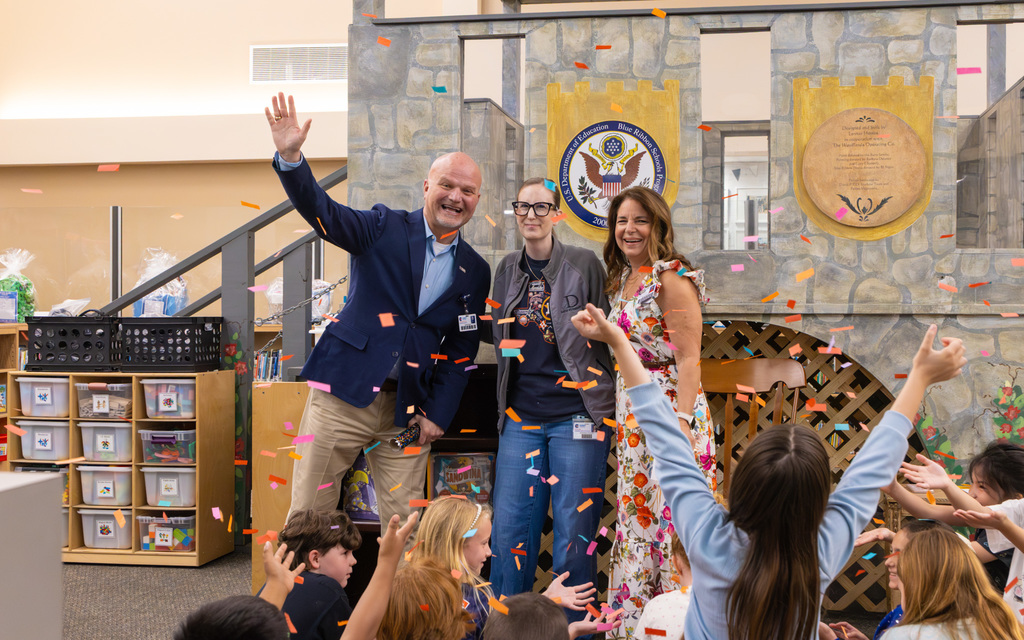 Three adults and children share a lively celebration with confetti raining down in a classroom-like setting to recognize a teacher