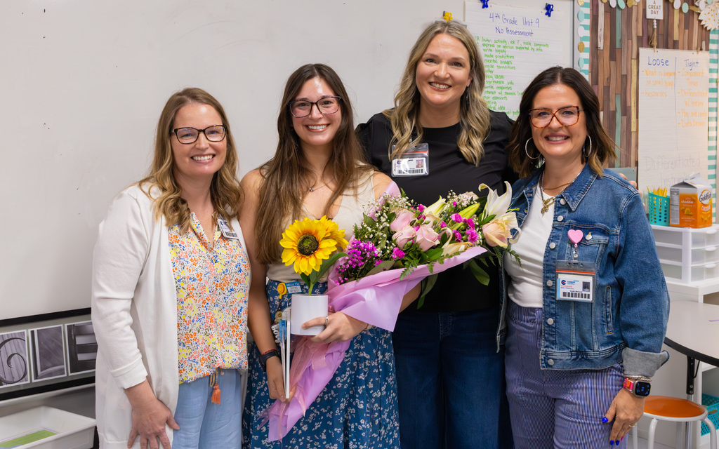 Four women smiling in a classroom setting, one holding a mixed bouquet and a sunflower in a small vase