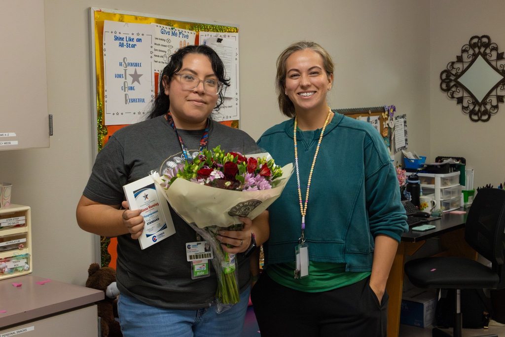 Staff member with colleague holding award plaque and flowers.