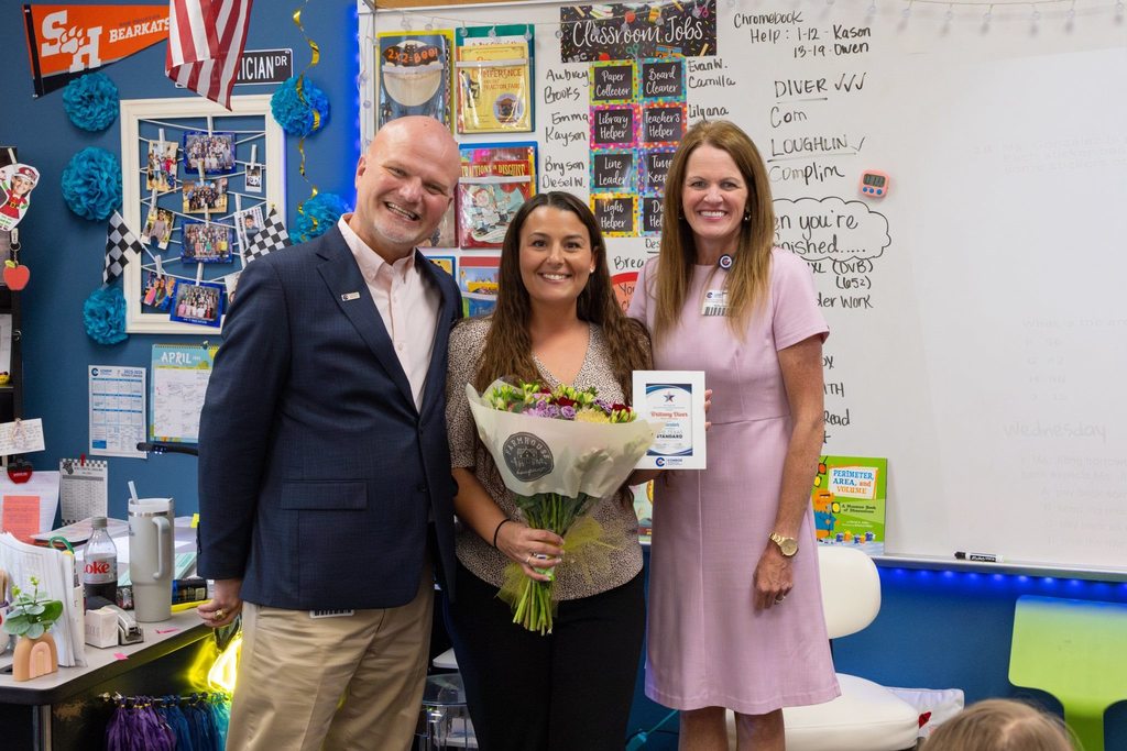 Superintendent and board member with teacher holding award plaque and flowers.