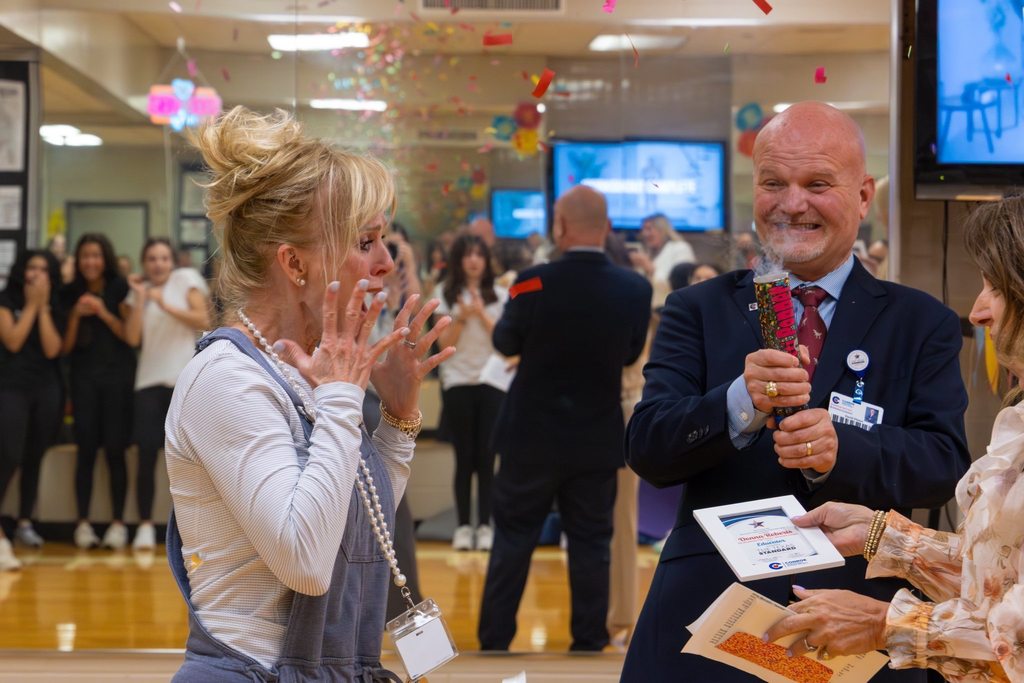 Staff member surprised with award plaque by board member while superintendent pops confetti.