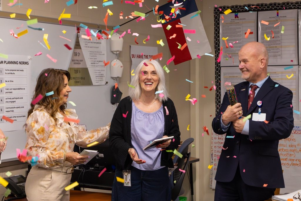Staff member holding award plaque congratulated by Board President while Superintendent pops confetti.
