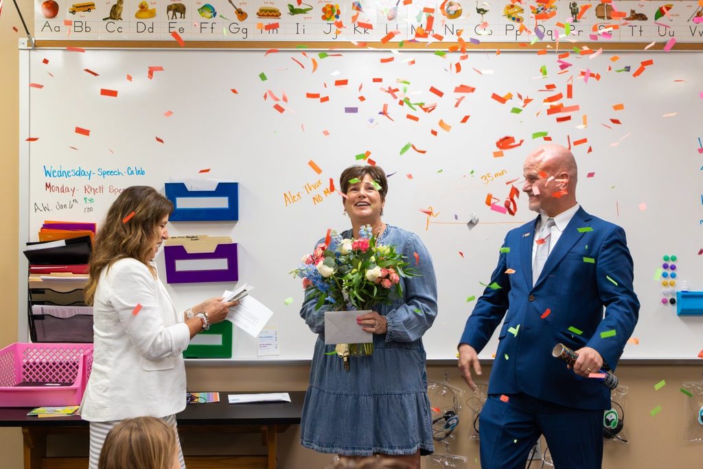 Superintendent and Board President present staff member holding flowers with award as confetti falls.