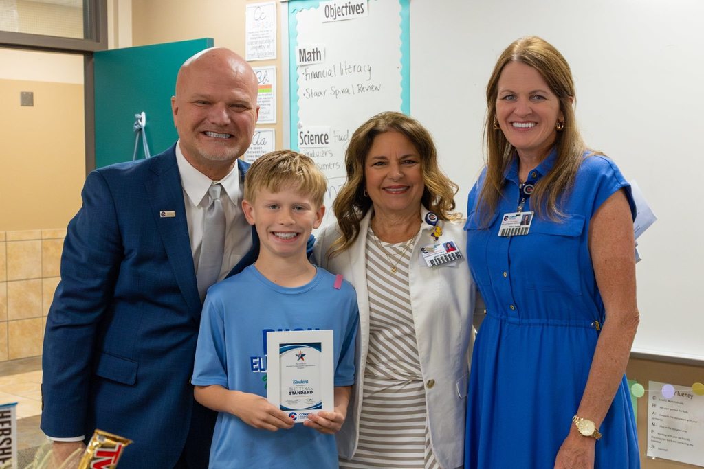 Superintendent and two board members congratulate student holding award plaque.