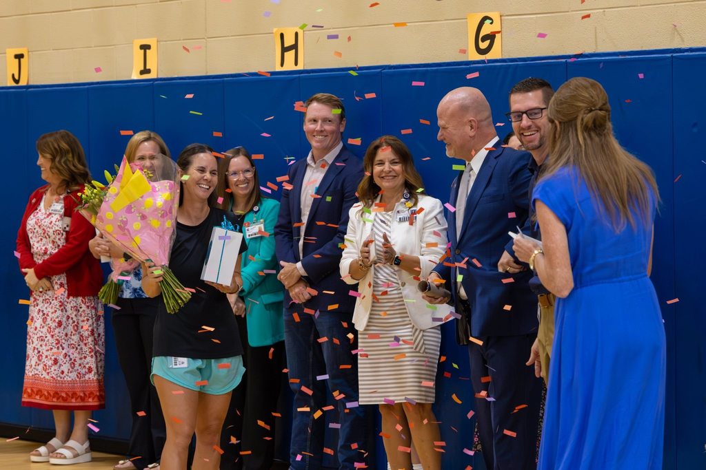 Staff member holding flowers and award congratulated by administration and family as confetti falls.
