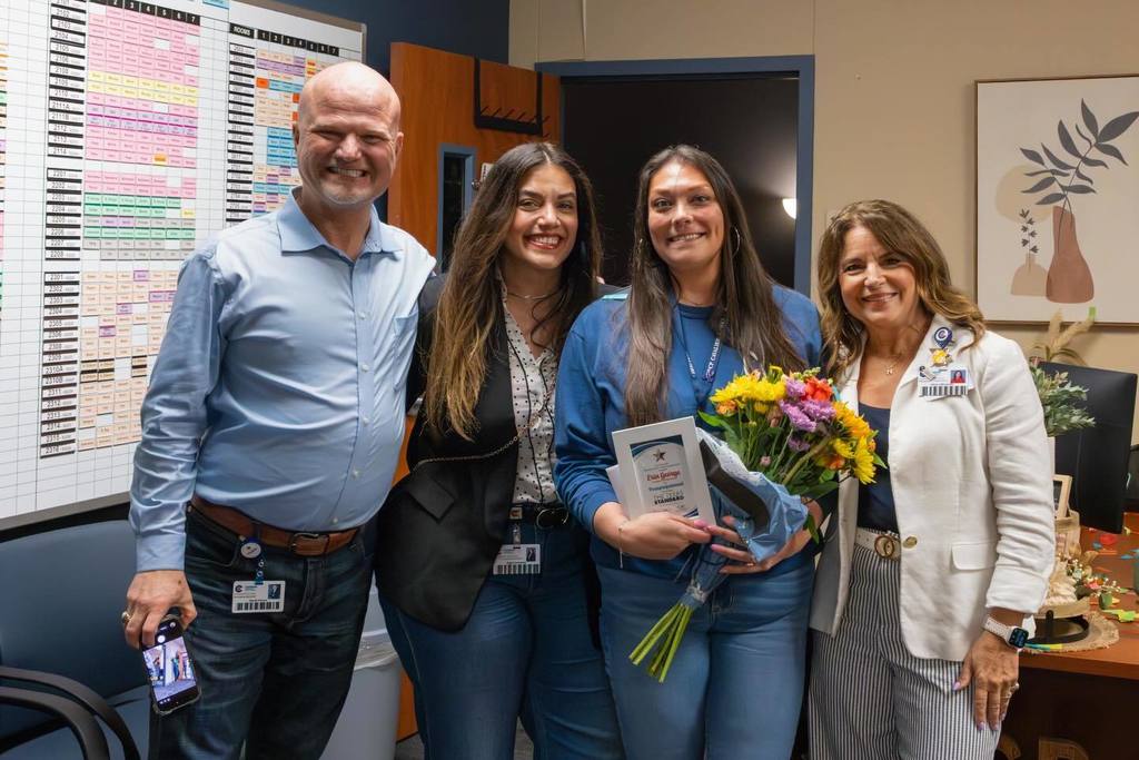 Superintendent and two board members congratulate staff member holding award plaque and flowers.