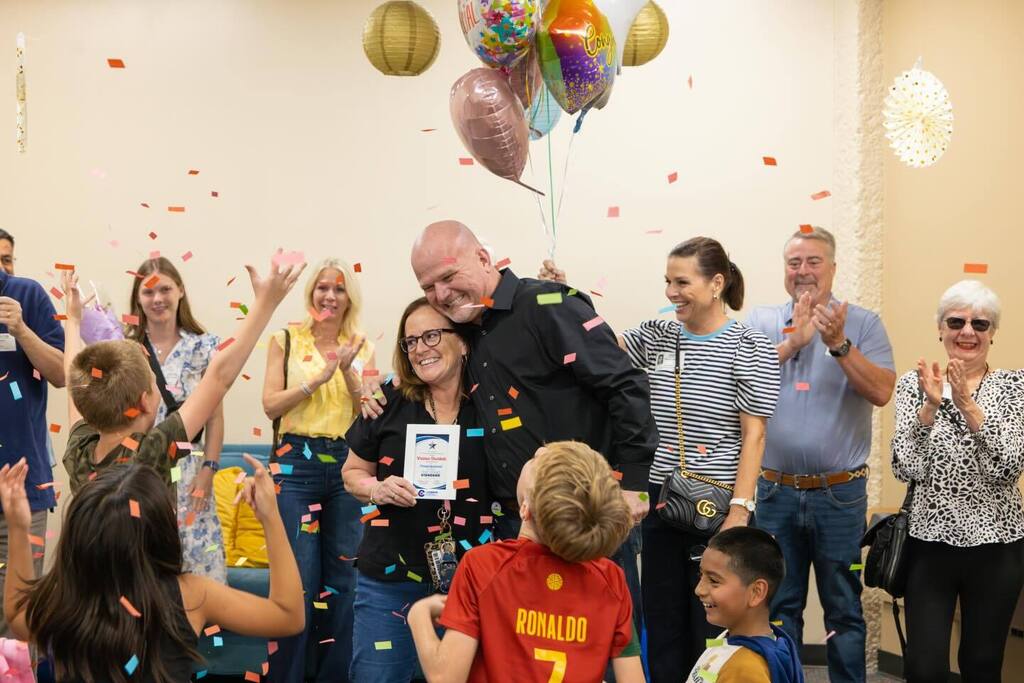 Staff member holds award plaque and balloons and hugs Superintendent as confetti falls.